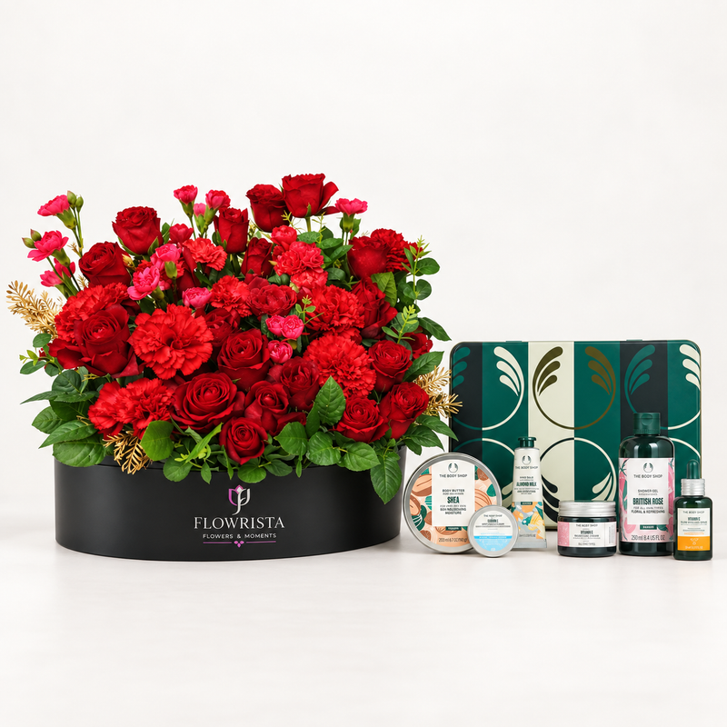 A curated gift set featuring a large black round flower box filled with red roses, carnations, and green foliage, alongside bath and body items including a decorative tin, a round jar, and three bottles from Flowrista. The Flowrista brand is clearly visible on the flower box and product labels.