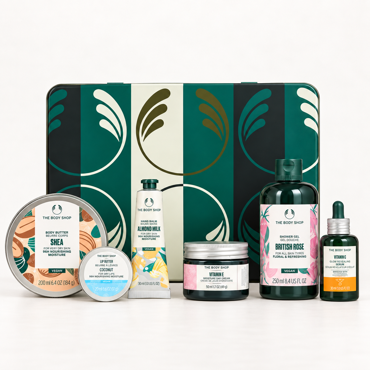 The Body Shop Bath & Body Set featuring a green and cream patterned gift tin with six bath products arranged in front on a white background: Shea butter, Almond Milk hand wash, British Rose shower gel, Hydrate body wash, and two Vitamin dropper bottles.