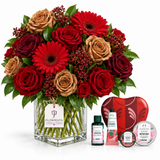 A large floral arrangement in a clear square glass vase with red and tan roses, red gerbera daisies, green leaves, and small red berry clusters, displayed with a Flowrista tag. Beside the flowers are small gift items including a red heart-shaped box, a pink bottle, a pink tube, and a round tin labeled Strawberry Shortcake, all on a white background.