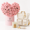 A curated gift set featuring a heart-shaped arrangement of pink roses in a pink cylindrical box by Flowrista, a cream monogram backpack with gold hardware, a matching cream candle, and a small cream pouch with gold keychain charms, all arranged on a clean white background.