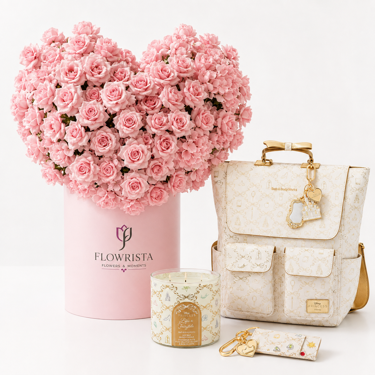 A curated gift set featuring a heart-shaped arrangement of pink roses in a pink cylindrical box by Flowrista, a cream monogram backpack with gold hardware, a matching cream candle, and a small cream pouch with gold keychain charms, all arranged on a clean white background.