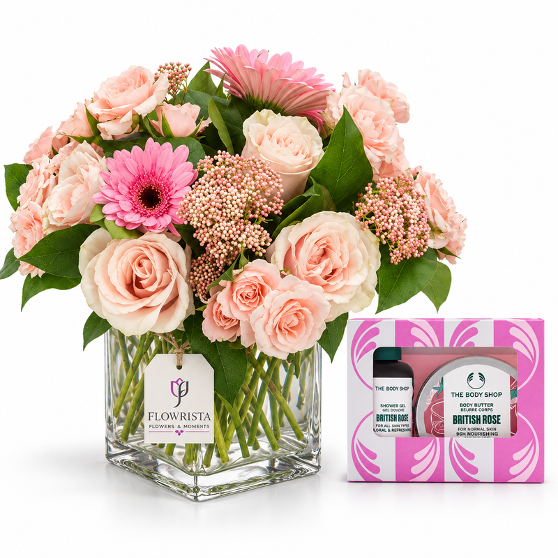 A floral arrangement in a clear square glass vase with pink and blush roses, pink gerbera daisies, and green foliage, displayed beside a pink and white The Body Shop Body Butter gift box on a white background.