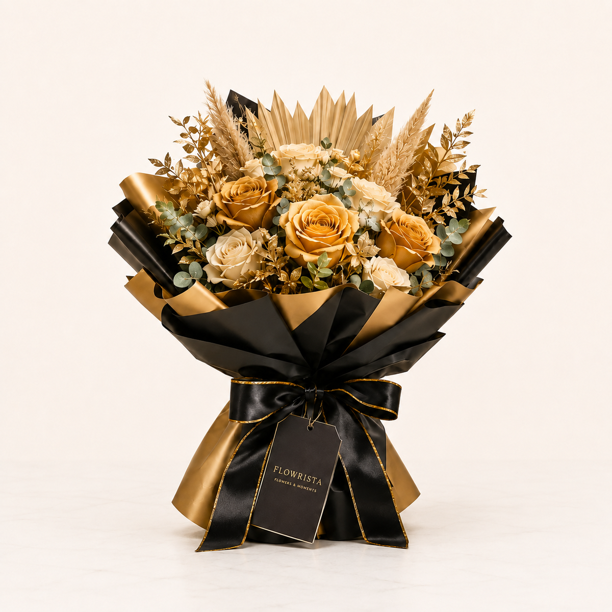 A dried flower bouquet in black and gold wrapping with a ribbon bow and tag, featuring golden roses, beige filler flowers, and dried grasses on a clean off-white background.