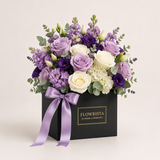 A floral bouquet in a black square box with a lavender ribbon and a gold-embossed 'FLOWRISTA FLOWERS & MOMENTS' label. The bouquet features purple and white roses, hydrangeas, lavender sprigs, and green foliage on a light beige background.