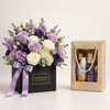 A floral gift box arrangement with purple and white flowers, green foliage, and a lavender ribbon, next to a beige gift bag containing bath and body items. The flower box is labeled FLOWWRISTA FLOWERS & FRAGRANCE.