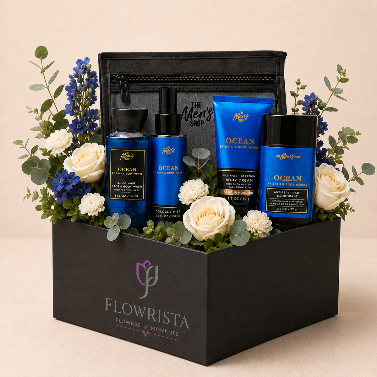 A curated men's self-care gift set arranged in a black rectangular box with the brand name FLOWRISTA visible on the front. The set includes blue and black bottles and tubes from The Men's Shop, including Ocean Body Wash, Ocean Body Lotion, and Ocean Body Cream, styled with white roses, blue flowers, and green foliage against a warm beige background.