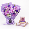 A purple and lavender flower bouquet wrapped in matching purple paper, tied with a ribbon, and displayed next to a pink candle on a decorative pink cushion against a white background.
