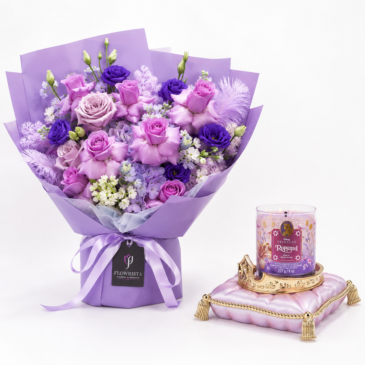 A purple and lavender flower bouquet wrapped in matching purple paper, tied with a ribbon, and displayed next to a pink candle on a decorative pink cushion against a white background.