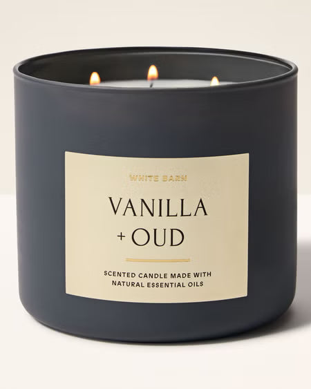 A matte black three-wick scented candle labeled 'White Barn Vanilla + Oud' with three lit flames, set against a clean off-white background.