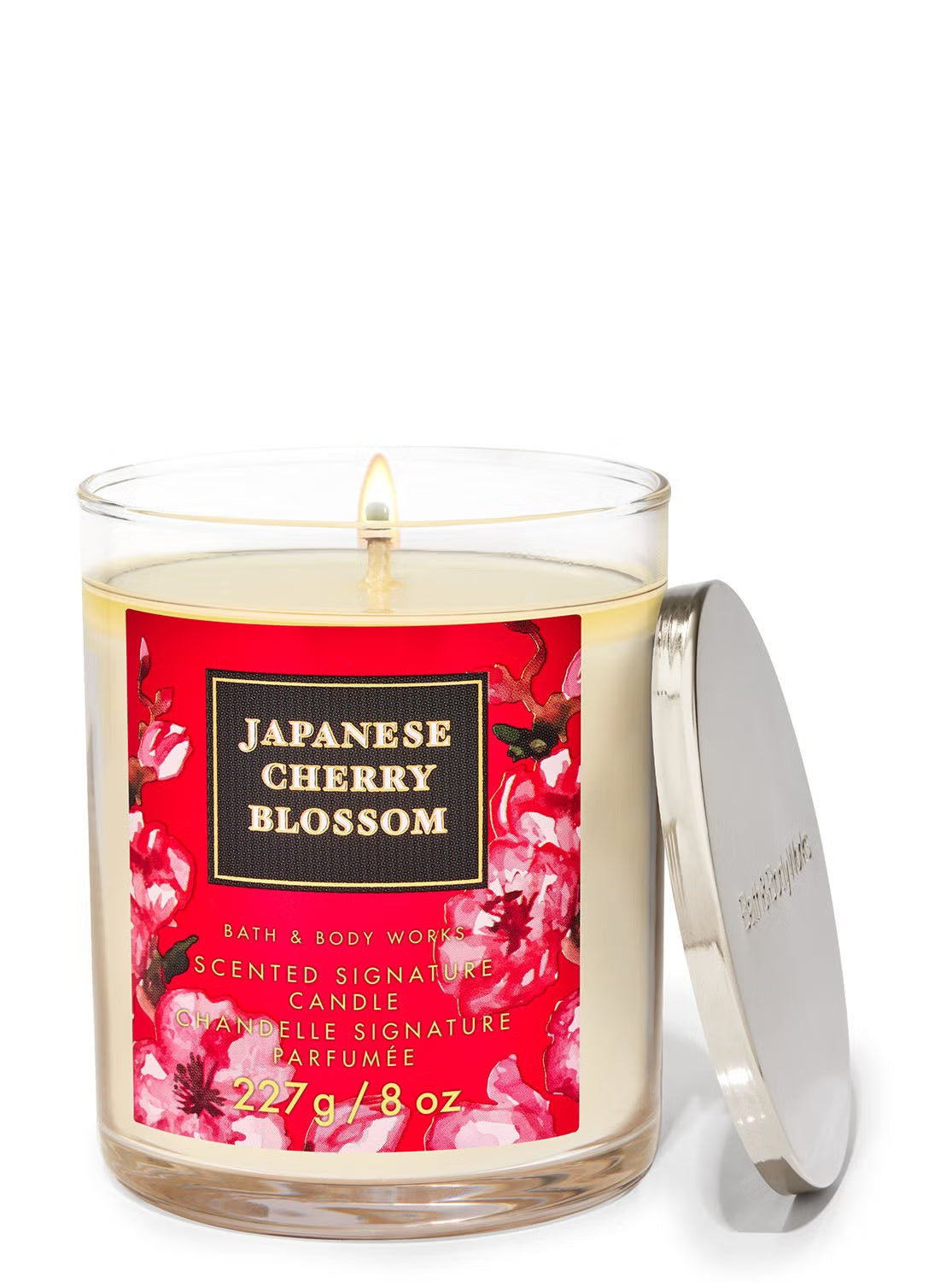 A Bath & Body Works Japanese Cherry Blossom scented candle in a clear glass jar with a red floral label, shown with its silver metal lid leaning against the right side on a white background.