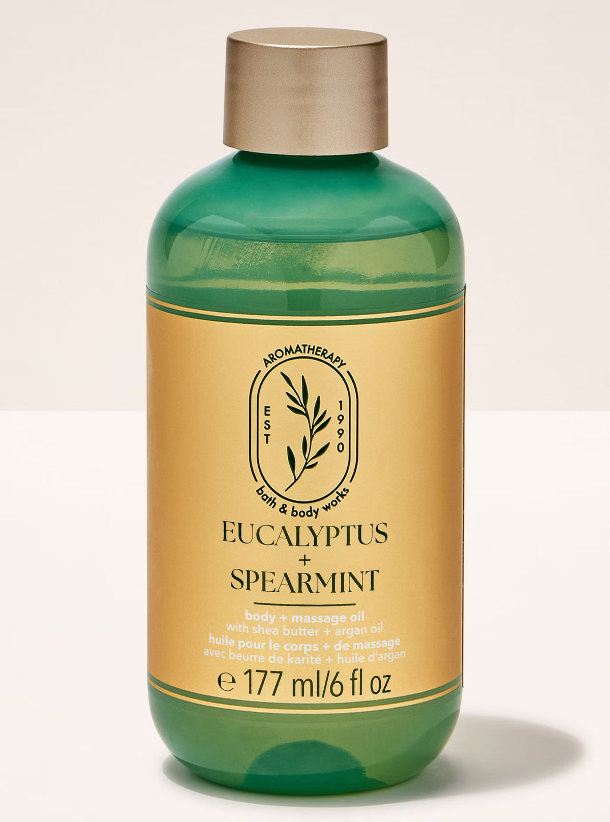 A green translucent bottle of Bath & Body Works Eucalyptus + Spearmint massage oil with a gold cap and a gold label, set against a light beige background.