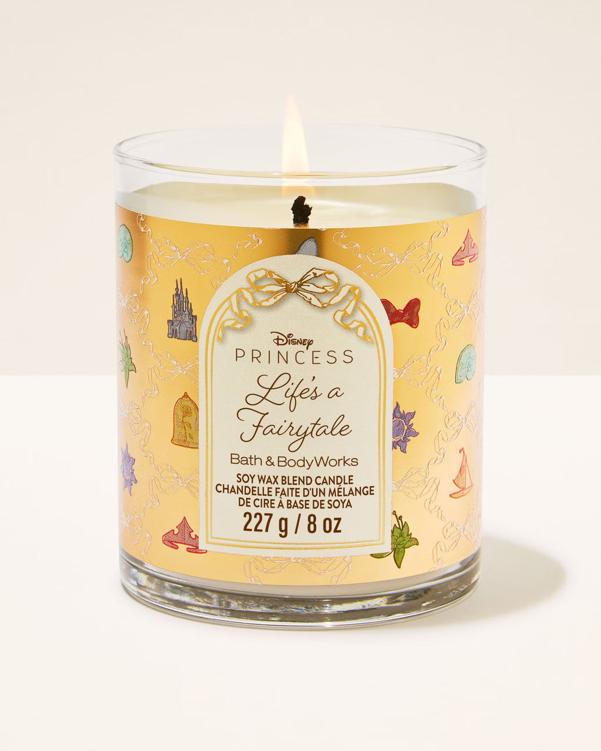 A Disney Princess 'Life's a Fairytale' scented candle in a clear glass jar with a yellow label decorated with fairytale castle and bird illustrations. The candle is lit, showing a small flame, and is photographed against a clean, off-white background.