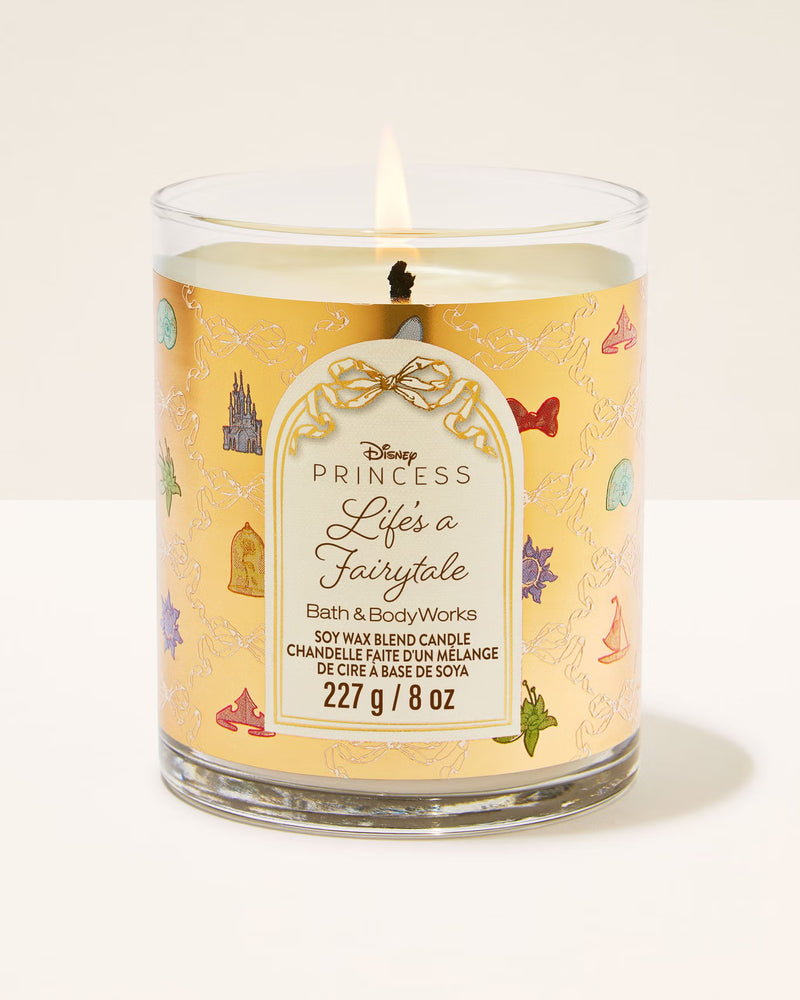A Disney Princess 'Life's a Fairytale' scented candle in a clear glass jar with a yellow label decorated with fairytale castle and bird illustrations. The candle is lit, showing a small flame, and is photographed against a clean, off-white background.