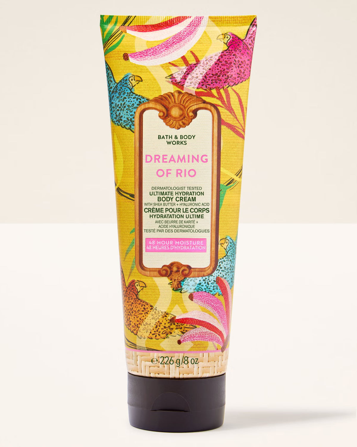 A squeeze tube of Bath & Body Works Dreaming of Rio Ultimate Hydration Body Cream, featuring a vibrant yellow background with colorful tropical bird and floral illustrations, set against a plain off-white background.