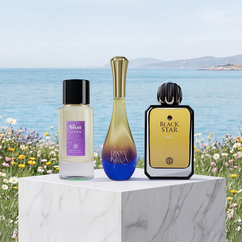 Three perfume bottles displayed on a white marble pedestal outdoors by the sea, with flowers in the foreground. The bottles are labeled Viveri La Rose, Annalena, and Black Star.