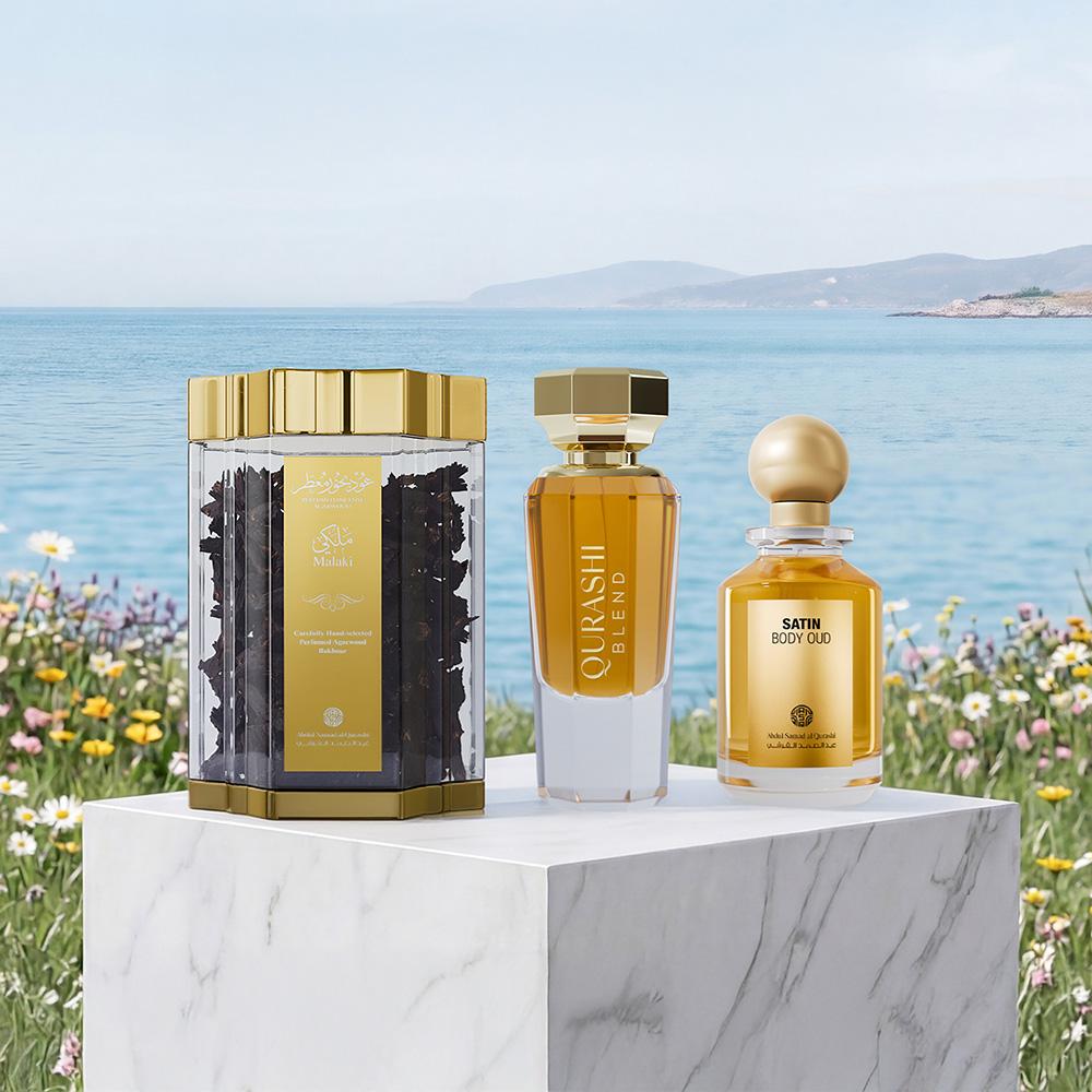 Three fragrance products from QuraSh Blend are displayed on a white marble pedestal with a scenic beach and wildflower background. The set includes a clear hexagonal tin filled with dark incense sticks, a tall rectangular glass bottle with amber liquid and gold detailing, and a round glass bottle with amber liquid and a gold spherical cap.