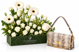 A luxury gift set featuring a green rectangular box of white flowers, including daisies and roses, next to a beige snakeskin-patterned shoulder bag with gold chain and lock details. The box is branded 'FLOWRISTA'.