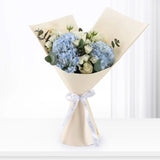 A wrapped flower bouquet with light blue hydrangeas, white roses, and green eucalyptus stems, wrapped in beige paper with a white ribbon tied in a bow, set against a bright white background.
