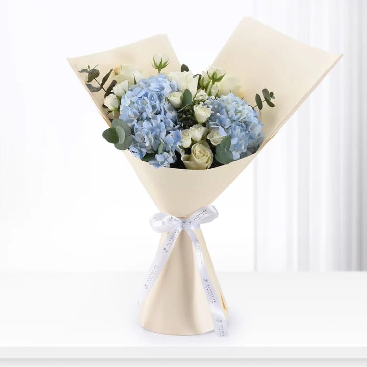 A wrapped flower bouquet with light blue hydrangeas, white roses, and green eucalyptus stems, wrapped in beige paper with a white ribbon tied in a bow, set against a bright white background.