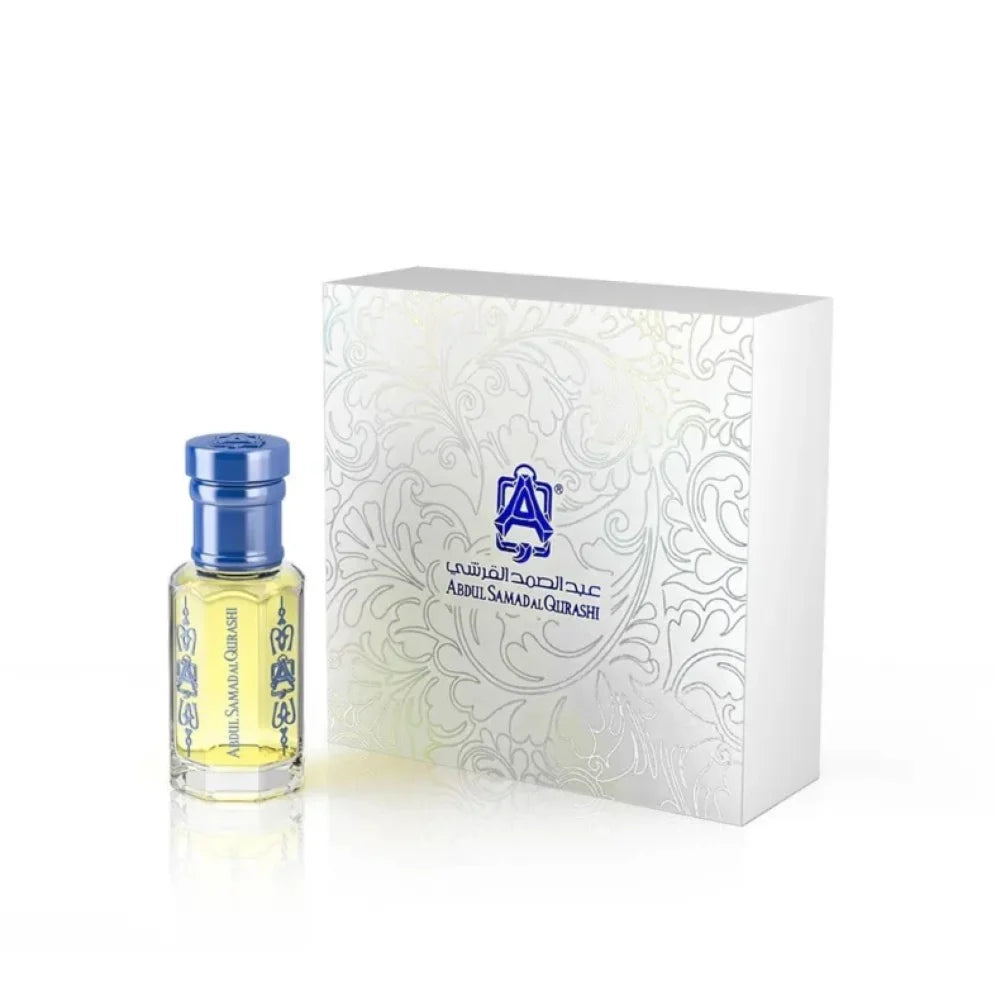 A small clear glass perfume bottle with pale yellow liquid and a metallic blue cap stands beside a white rectangular presentation box with embossed floral detailing and blue branding text.