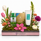 A floral gift basket arrangement by Flowrista featuring a green and white facial mask care set, a gold box, and a white tube, styled with tulips, chrysanthemums, and an orchid in a pink and gray tray on a white background.