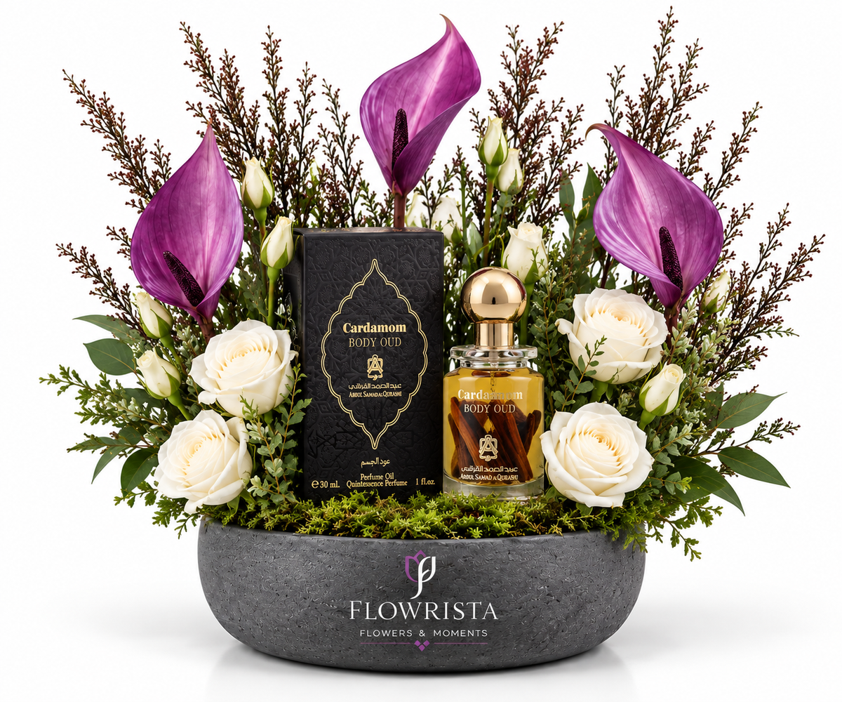 A luxury floral gift arrangement by Flowrista featuring a bottle of Cardamom EDP and its black box nestled in a bowl of white roses, purple calla lilies, green foliage, and moss, set against a solid white background.