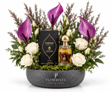A luxury floral gift arrangement by Flowrista featuring a bottle of Cardamom EDP and its black box nestled in a bowl of white roses, purple calla lilies, green foliage, and moss, set against a solid white background.