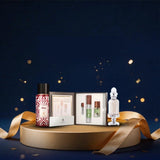 A luxury gift set displayed on a round gold pedestal against a dark navy blue background with gold bokeh and ribbon accents. The set includes a patterned body wash bottle, an open box with three small cosmetic tubes, and a clear perfume bottle, all arranged with draped gold ribbons.
