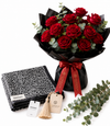 A bouquet of 18 red roses wrapped in black paper with a red ribbon, displayed alongside a black-and-white patterned scarf, a folded black garment, and eucalyptus stems on a white background. The tags show the brand Mango.