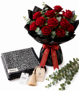 A bouquet of 18 red roses wrapped in black paper with a red ribbon, displayed alongside a black-and-white patterned scarf, a folded black garment, and eucalyptus stems on a white background. The tags show the brand Mango.