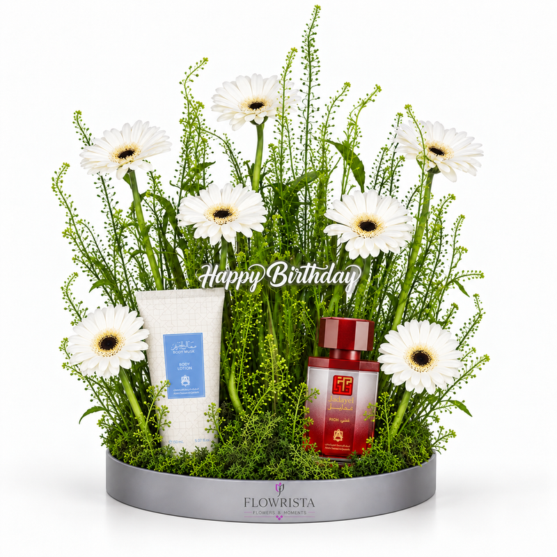 A floral gift arrangement in a round silver tray, featuring white daisies, green foliage, a white tube product with a blue label, and a red cosmetic bottle. The arrangement is set against a plain white background with a 'Happy Birthday' text overlay.