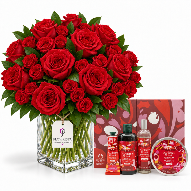 A large bouquet of red roses in a clear glass vase with green stems, displayed with matching red gift items including a box, hand cream tubes, a bottle, and a tin on a white background. The tag on the vase reads Flowrista.