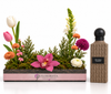 A floral arrangement in a rectangular pink and dark gray planter by Flowrista, displayed next to a textured brown bottle labeled 'Grey Pearl Diamonds' on a clean white background.