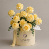 A round cream-colored gift box filled with pale yellow roses and green eucalyptus stems, with a cream tassel and white tissue paper. The box is branded with the logo and name 'FLOWRISTA'.