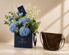 A luxury gift set from Flowwrista featuring a floral arrangement with blue hydrangeas, blue tulips, and green foliage in a navy blue branded box, alongside a brown suede shoulder bag on a beige marble surface.
