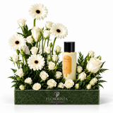 A floral gift arrangement in a dark green rectangular box from Flowrista, featuring white roses, white gerbera daisies, and green foliage, with a bottle of body wash placed among the flowers on a white background.