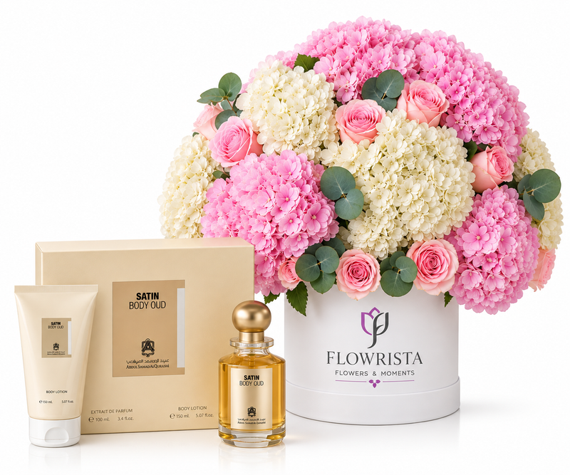 A luxury gift set featuring a large floral arrangement of pink and white flowers in a white bucket labeled 'FLOWRISTA', alongside a beige box, a tube of body lotion, and a small bottle of satin body ood. The products are arranged on a clean white background.
