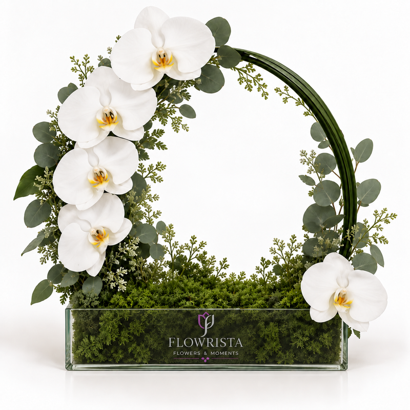 A floral arrangement in a rectangular glass planter by Flowrista, featuring white orchids, green eucalyptus, and moss inside a curved green wire frame, photographed on a white background.
