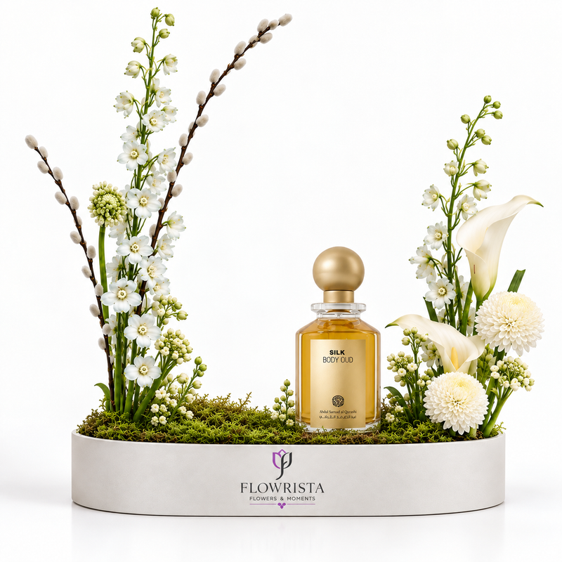 A rectangular white planter with the brand name FLOWWRISTA, filled with green moss and decorated with white flowers, tall green stems, and a central gold perfume bottle labeled E.L.