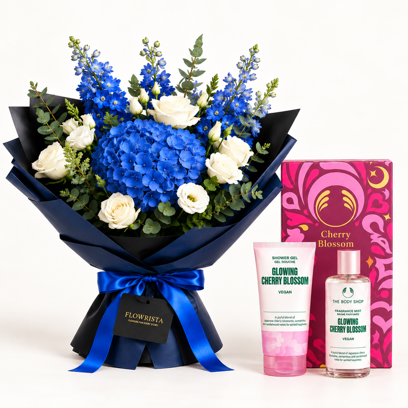 A gift set featuring a large bouquet of blue hydrangeas, white roses, and blue delphiniums in dark blue wrapping with a blue ribbon, alongside a pink box labeled Cherry Blossom and two skincare products from The Body Shop: Glow-ing Cherry Blossom body gel and shower gel.