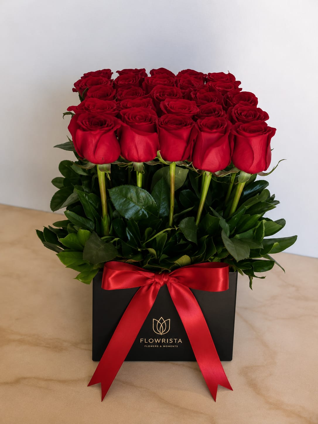 A box of 20 red roses with green stems and leaves, presented in a black square box tied with a red satin ribbon. The box features the brand name 'FLOWRISTA' printed in gold.