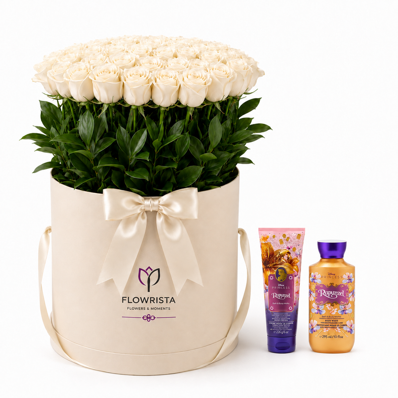 A large round cream gift box filled with white roses and green foliage, tied with a cream satin bow. The box is branded FLOWWRISTA and stands next to two bath/body products from the same brand on a clean white background.