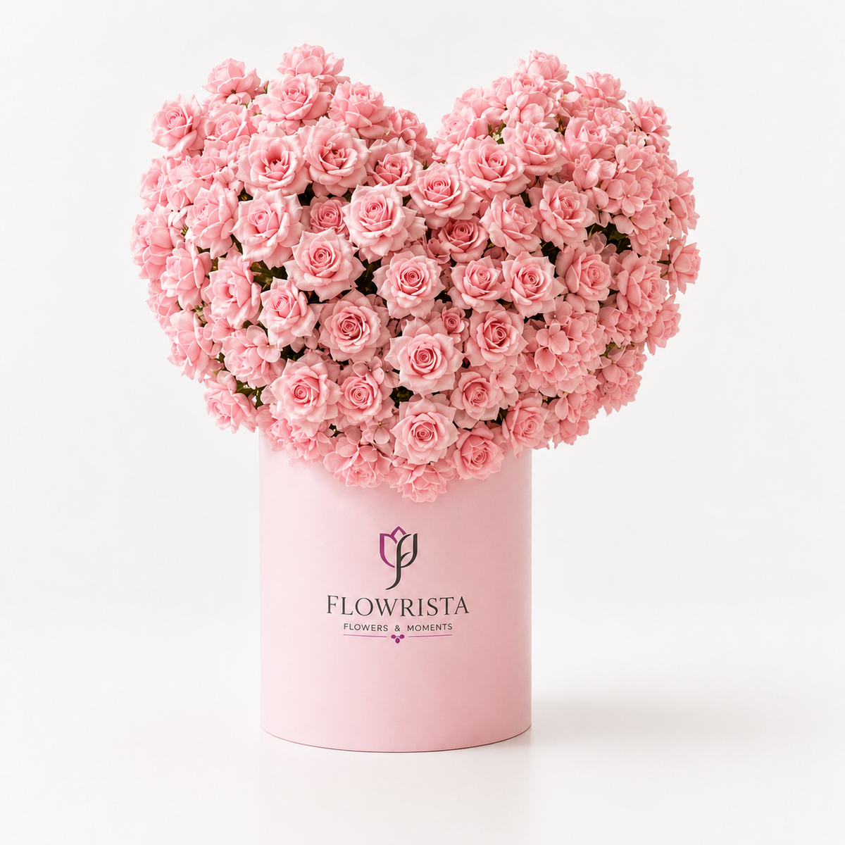 A heart-shaped arrangement of light pink roses in a cylindrical pink box with the brand name FLOWRISTA printed on the front, set against a clean white background.