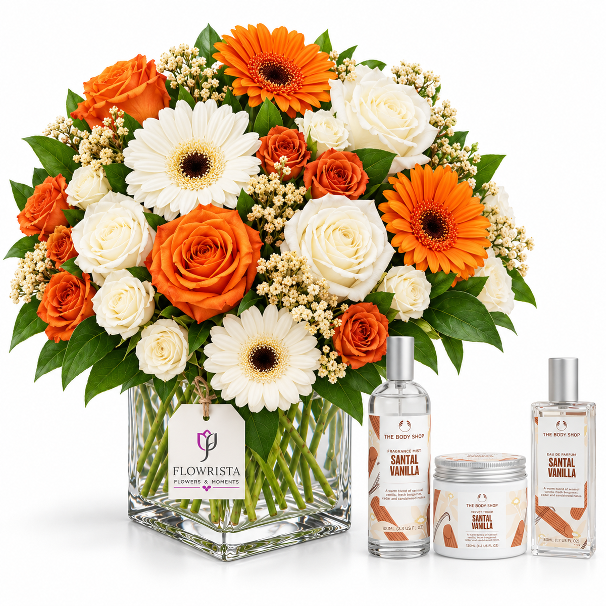 A floral arrangement in a clear square glass vase with orange gerbera daisies, white roses, orange roses, and green foliage, displayed beside three matching Flowrista Santa Vanilla scented products on a white background.