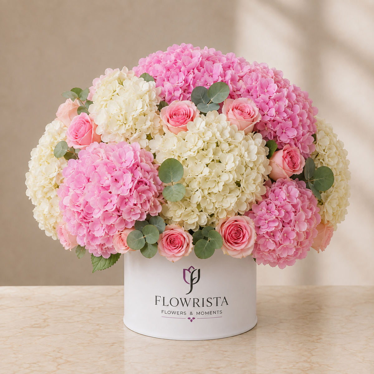 A round white box filled with a floral arrangement of pink and cream hydrangeas, pink roses, and green eucalyptus leaves, displayed on a beige marble surface against a neutral beige background. The box is branded with 'FLOWRISTA FLOWERS & MOMENTS'.
