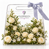 Creamy white roses and small white flowers in a clear Flowrista acrylic handled bag with lavender bow