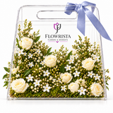 Fresh white roses and filler flowers in a transparent handled Flowrista gift bag with lavender ribbon