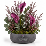 Artificial purple calla-style flowers with burgundy sprigs and eucalyptus-style greenery in a textured grey Flowrista bowl