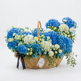 Fresh blue and white hydrangeas, white roses, spray roses and carnations in a woven Flowrista handled basket
