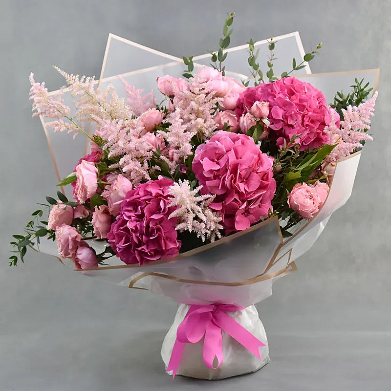 A vibrant bouquet of pink flowers, including hydrangeas, carnations, and pink astilbe, wrapped in white tissue paper with a pink ribbon and clear acrylic stand, set against a neutral gray background.